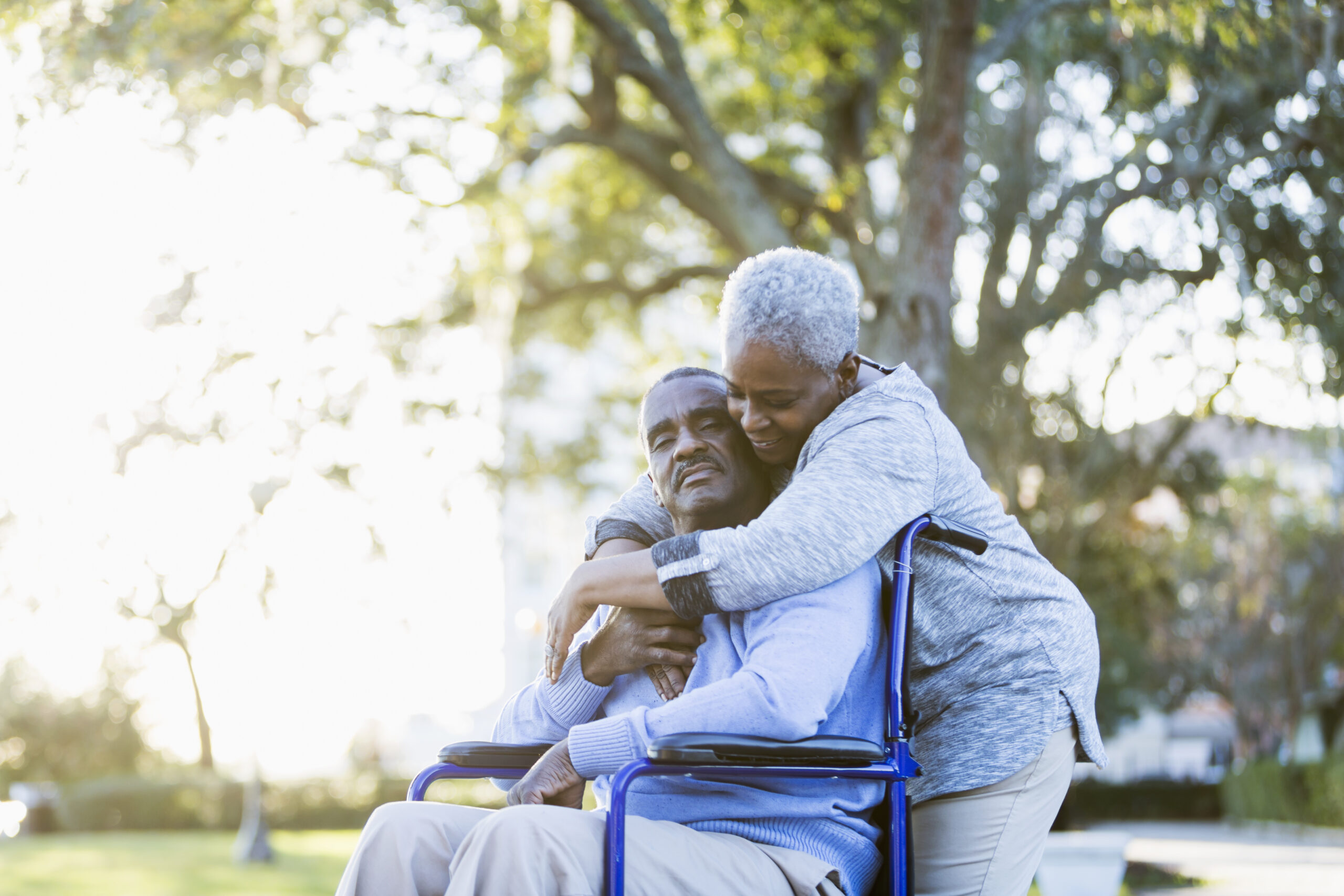 A woman standing behind a man’s wheelchair in a park leans down to hug him. When health issues force you off work, two types of long-term disability insurance can help.