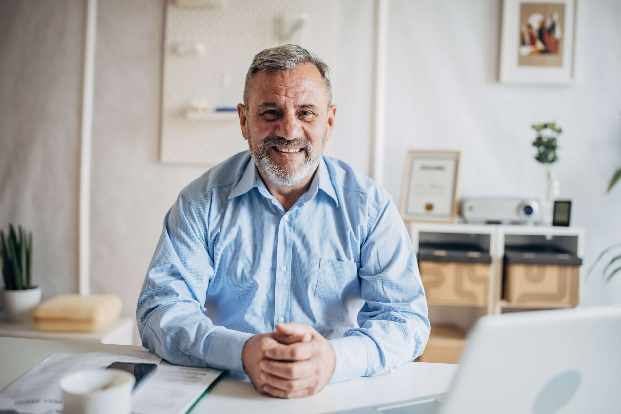 A man sits at a table over paperwork, clasping his hands and smiling for the camera. Barnes disability attorneys help you through the benefits process in Lehi, Utah.