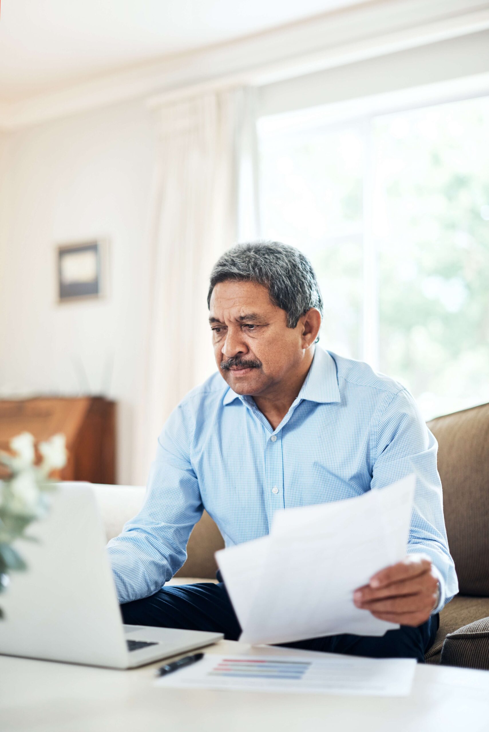 A man sits on his couch, holding papers and looking at a laptop. If you can’t work, options for help include Social Security Disability Insurance, Supplemental Security Income and long-term disability.