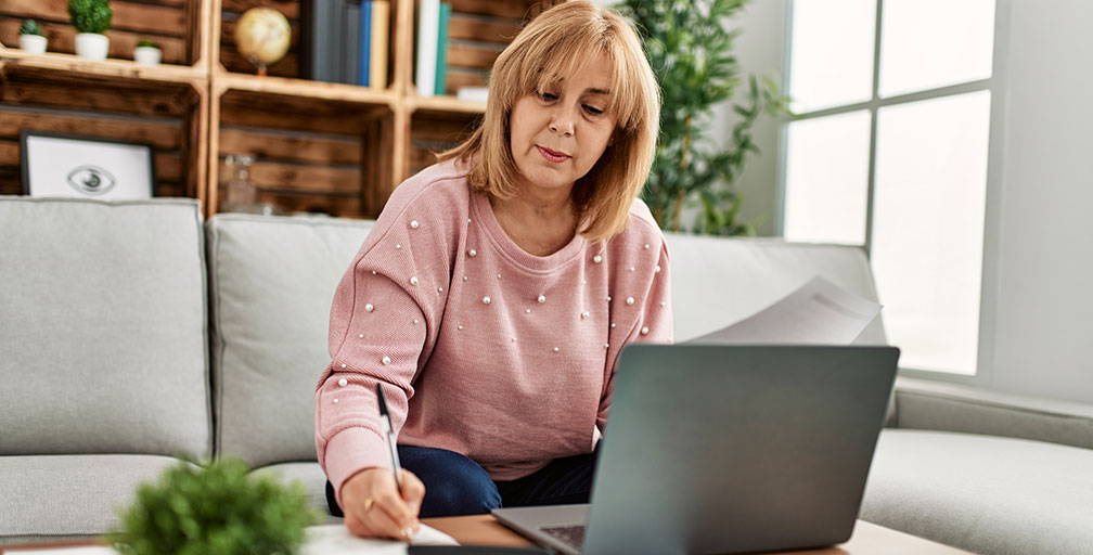 A woman sits on her couch writing on a piece of paper on the coffee table, laptop open. Barnes Disability Advocates help people file for disability benefits.