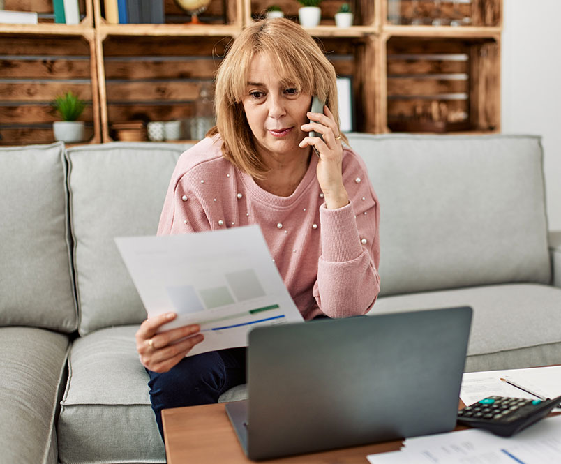 A woman sits on her couch talking on the phone, looking at a sheet of paper with her laptop open. Find out about different kinds of disability benefits you may qualify for from Barnes Disability lawyers in St. George.