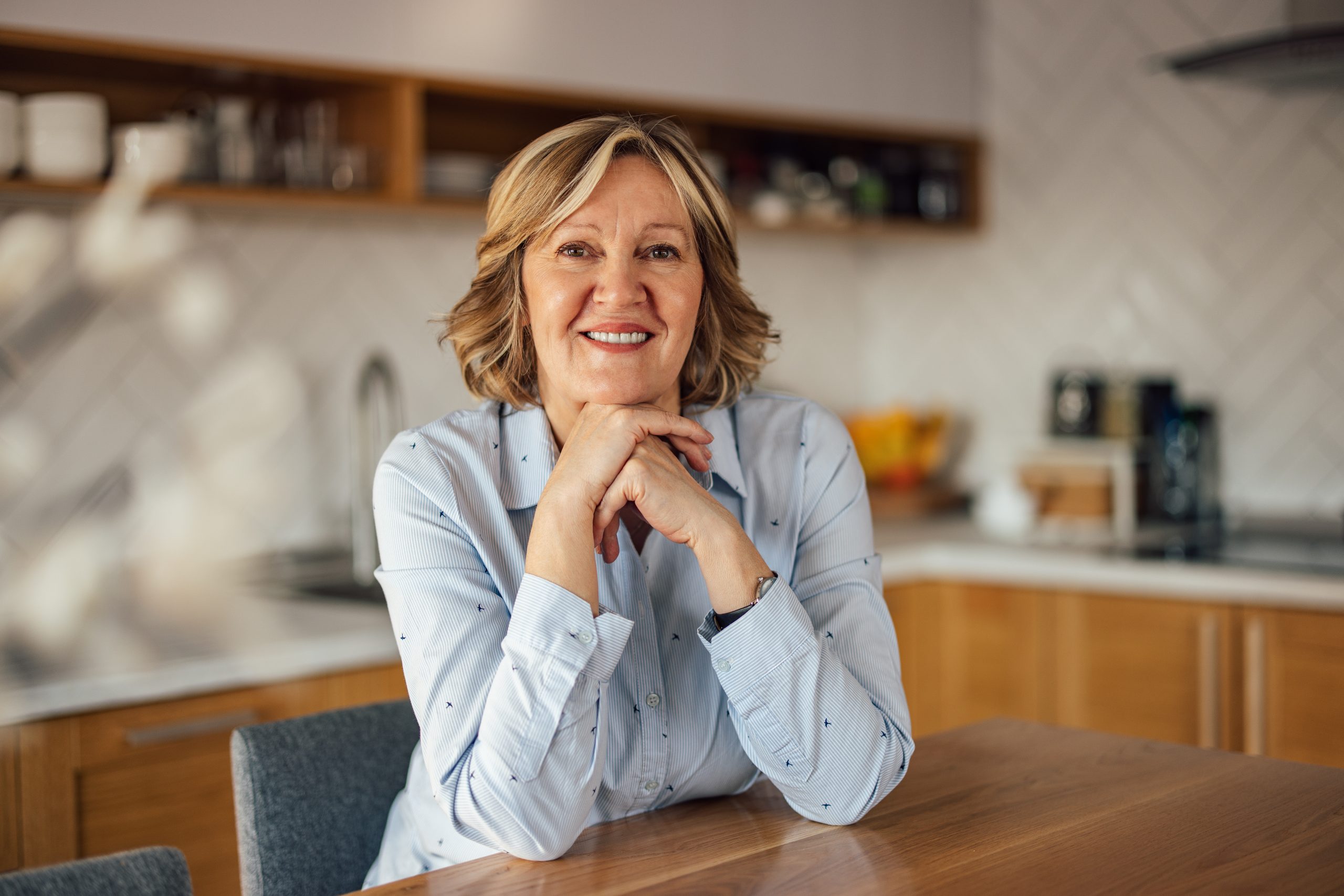 A woman sits at a kitchen table, elbows on the table, chin resting on hands, smiling at the camera. Past clients of Barnes Disability Advocates give satisfied reviews.